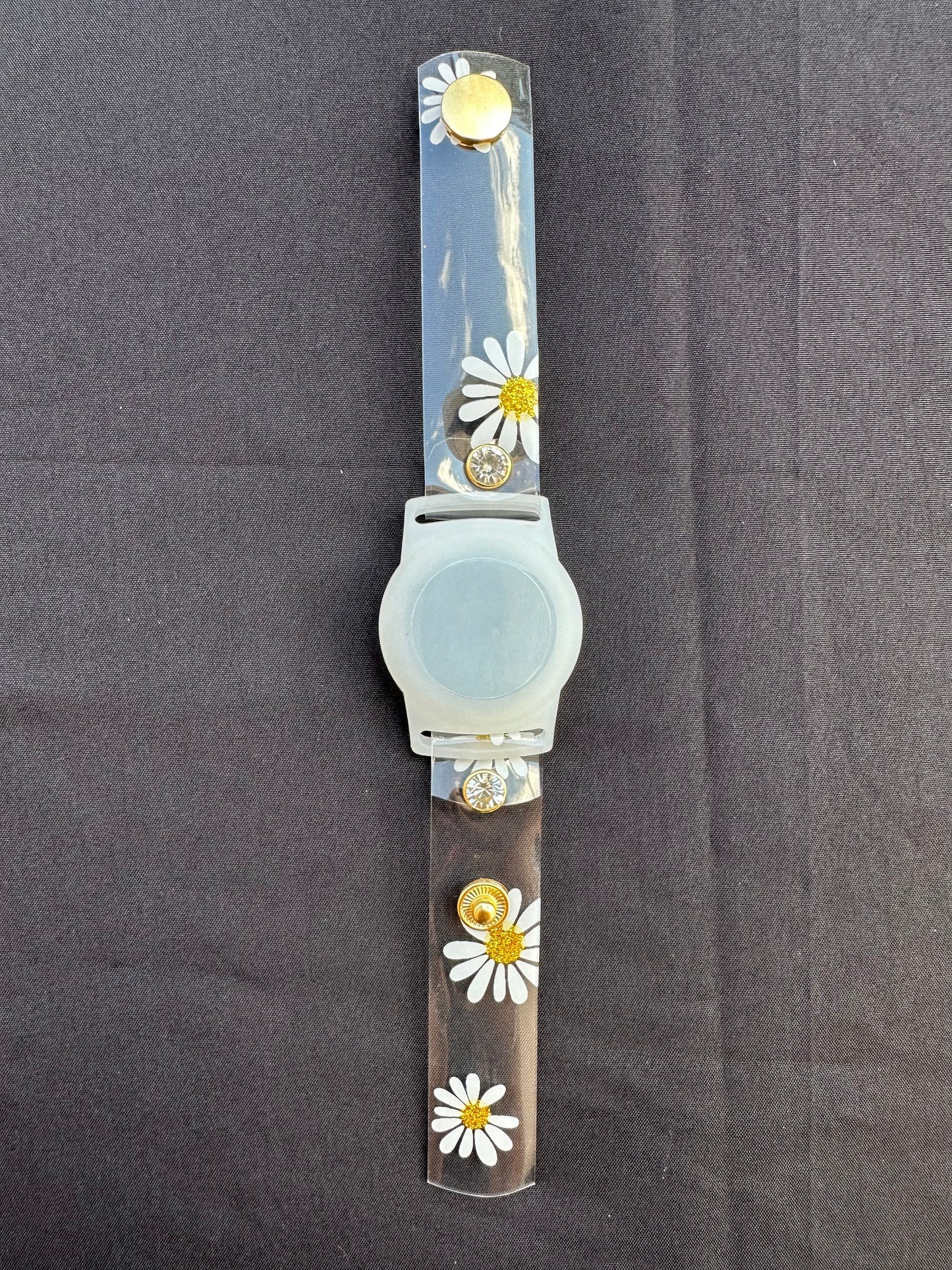 Anya style transparent PVC waterproof band perfect for all year round. The band has scattered daisies with a glittery gold center. The design is embellished with clear crystals with a gold rim. Frosted White Air Tag holder will act as the band's center.